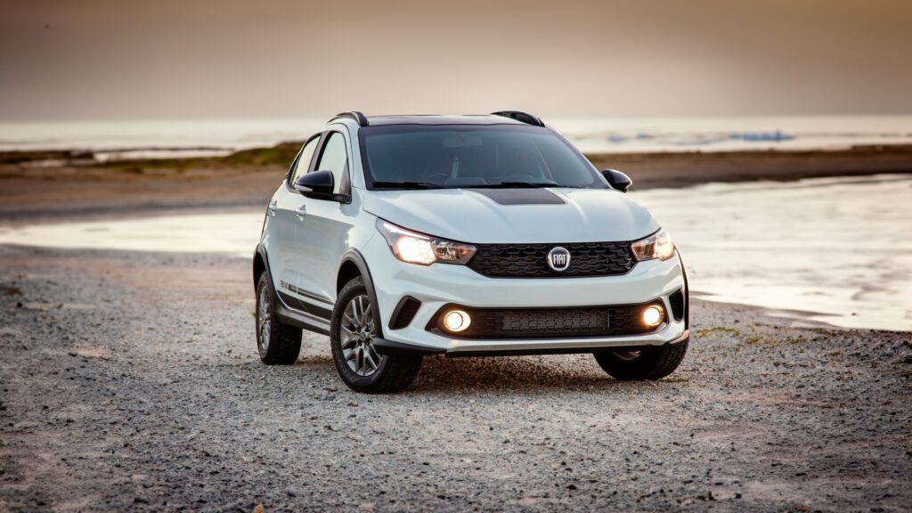 Imagem de divulgação do seguro do Fiat Argo. A imagem mostra um Fiat Argo estacionado em uma superfície de cascalho, com uma praia e o oceano ao fundo, durante o que parece ser o nascer ou o pôr do sol. O veículo está posicionado em um ângulo voltado para a câmera, destacando seu design frontal e perfil. O Fiat Argo possui uma grade proeminente com o logotipo da Fiat no centro, faróis elegantes e luzes de neblina integradas na área inferior do para-choque. A pintura do carro parece ser de uma cor clara, possivelmente branca ou prata, que contrasta com o teto de cor escura. As rodas têm um design de liga leve com vários raios.