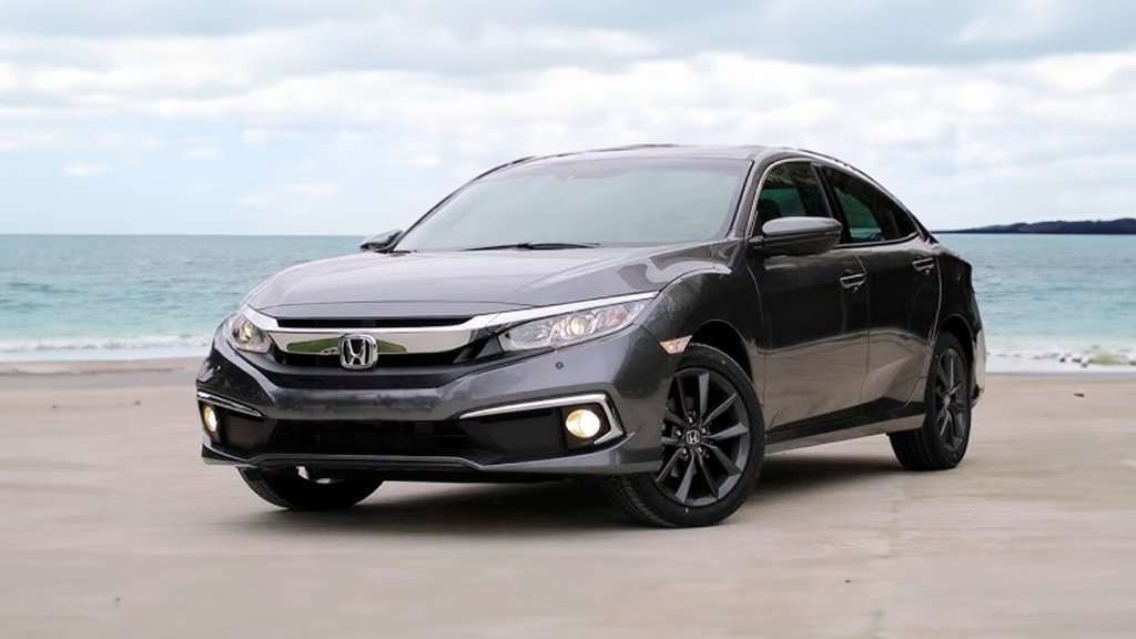 A imagem mostra um carro Honda Civic de cor cinza escuro. O carro está estacionado em uma praia, com o mar ao fundo e um céu com algumas nuvens. O Civic está posicionado de frente para a câmera, mostrando o seu design elegante e esportivo. Os faróis estão ligados, destacando as linhas modernas do veículo. As rodas de liga leve, com pneus largos, dão um ar mais robusto ao carro. A pintura cinza escuro confere um aspecto sofisticado ao Civic. No geral, a imagem transmite a sensação de um carro moderno e sofisticado, pronto para ser conduzido em um ambiente praiano e relaxante.