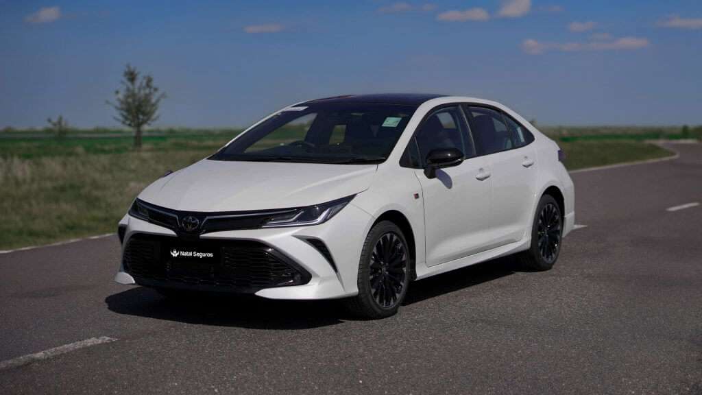 A imagem mostra um Toyota Corolla de cor branca, aparentemente um modelo mais recente. Ele está estacionado em uma estrada com uma leve curva, com um fundo de céu azul e algumas árvores. O carro está em um ângulo que permite uma visão frontal, destacando seu design elegante e moderno.