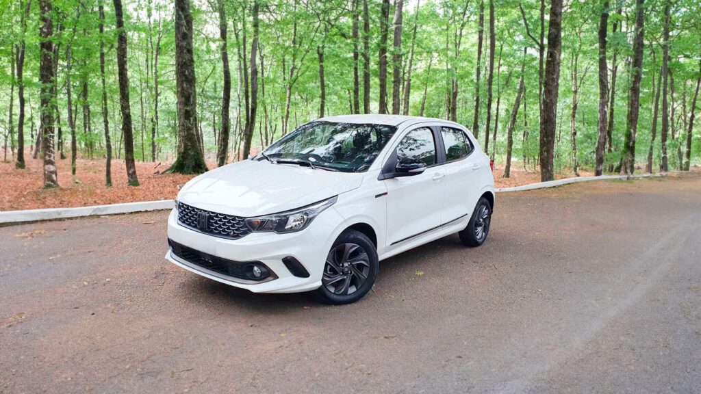A imagem mostra um Fiat Argo branco estacionado em uma estrada asfaltada, com uma floresta de árvores altas e verdes ao fundo. O veículo apresenta um design moderno com linhas elegantes, detalhes em preto na parte inferior da carroceria e rodas pretas. A grade frontal é proeminente, com um padrão de colmeia e o logotipo da Fiat no centro. Os faróis são afiados e angulares, contribuindo para a estética contemporânea do carro.