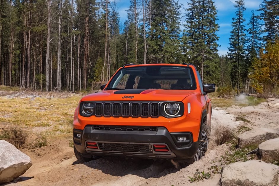 Imagem publicitária para o seguro do Jeep Renegade. A imagem mostra um Jeep Renegade Trailhawk estacionado em um ambiente ao ar livre, provavelmente em uma trilha ou terreno acidentado. O veículo é laranja com detalhes em preto e possui a grade de sete fendas característica dos veículos Jeep. Ao fundo, há altos pinheiros e um céu claro, sugerindo uma área remota ou selvagem. No chão, em frente ao veículo, há pedras, indicando que ele pode ser usado para condução off-road. A imagem destaca o design robusto do veículo e sua capacidade de enfrentar ambientes desafiadores, o que pode atrair aqueles interessados em aventuras e atividades off-road.