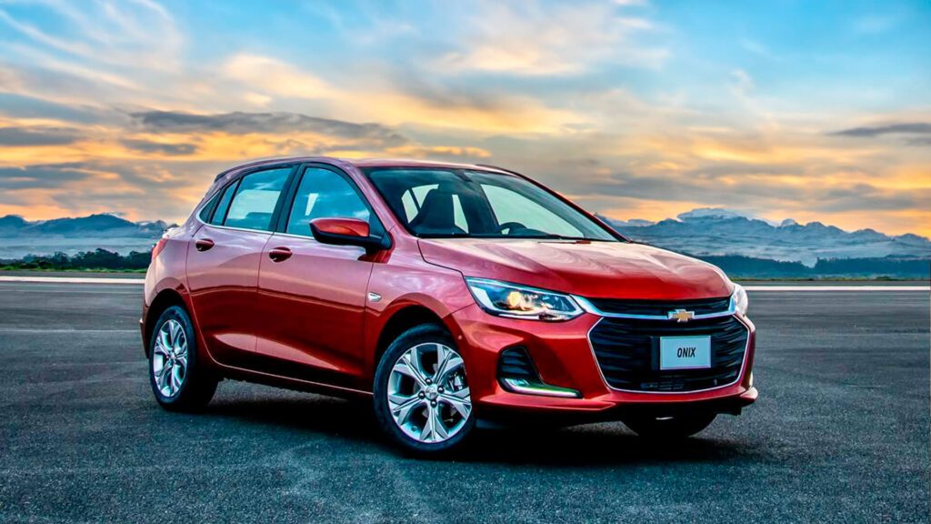 A imagem mostra um Chevrolet Onix hatchback vermelho estacionado em uma pista de pouso ao pôr do sol. O carro está de lado, com a frente voltada para a câmera. O sol está se pondo no horizonte, lançando uma luz laranja e dourada sobre o carro e a pista.