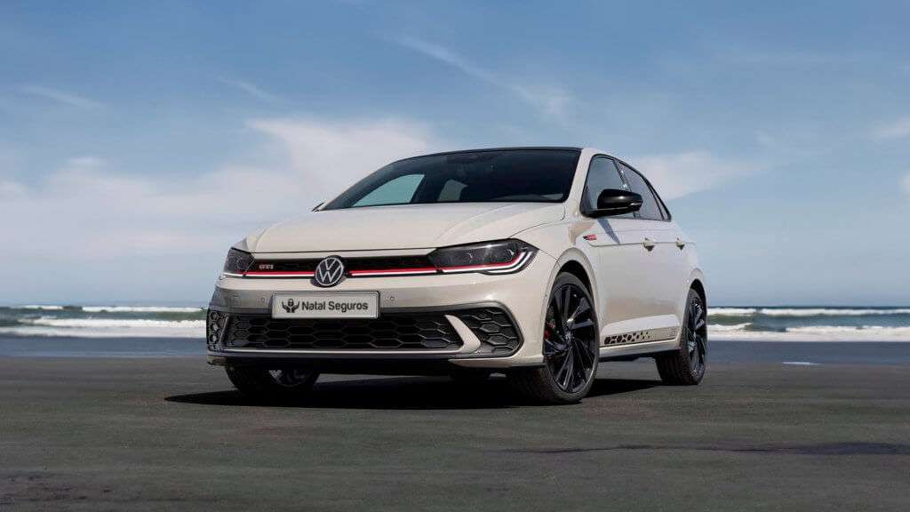 A imagem mostra um carro Volkswagen Polo GTI estacionado em uma área aberta, com o mar e o céu ao fundo. O carro é de cor branca e possui detalhes esportivos, como rodas pretas com acabamento brilhante, faróis de LED e uma grade frontal com um detalhe em vermelho. A placa do carro exibe o nome "Natal Seguros". O cenário de fundo inclui uma praia com ondas e um céu azul com algumas nuvens, sugerindo um dia claro e ensolarado. A imagem destaca o design esportivo e moderno do Polo GTI em um ambiente natural e agradável.