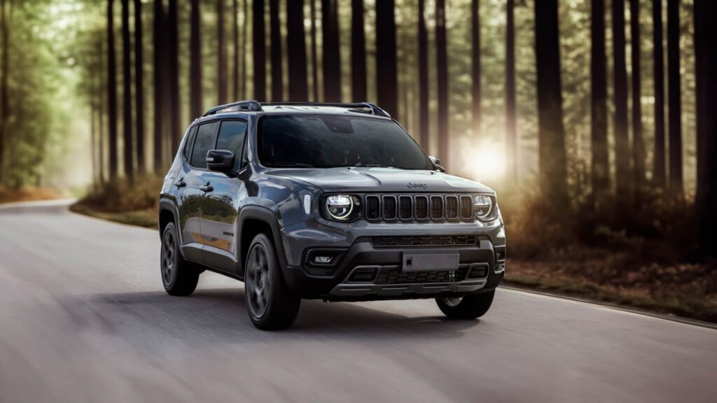 A imagem mostra o SUV Jeep dirigindo em uma estrada cercada por árvores altas, provavelmente em uma área florestal. O veículo está centralizado na imagem e é capturado de um ângulo frontal esquerdo, destacando características de design como a grade, os faróis e as rodas. O desfoque de movimento ao redor das rodas e do fundo sugere movimento, adicionando dinamismo à cena. Esta imagem pode ser relevante para mostrar o design do veículo ou para fins publicitários relacionados ao desempenho automotivo em ambientes naturais.