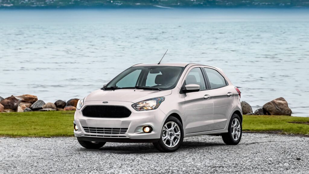 A imagem mostra um Ford Ka prateado estacionado em frente a uma praia ou lago. O cenário natural, com a água e as colinas ao fundo, cria uma cena interessante.