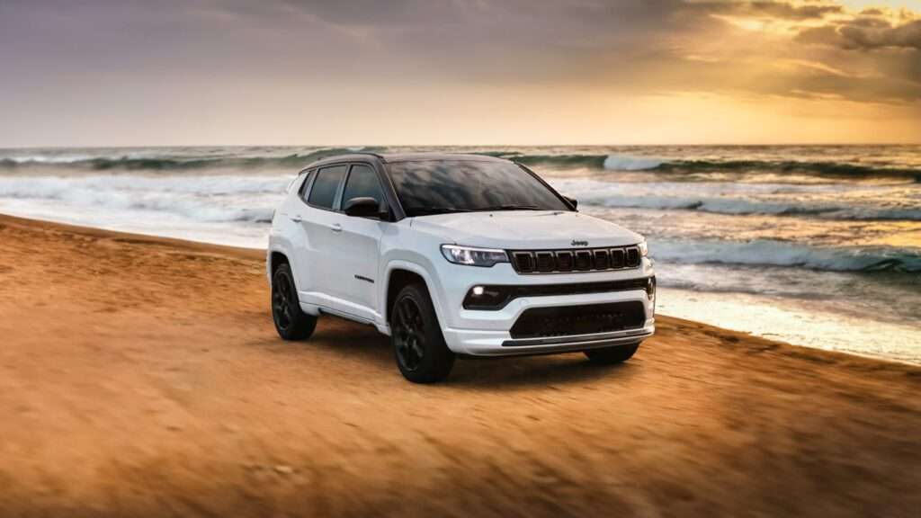 Imagem de divulgação do seguro do jeep compass. A imagem mostra um Jeep Compass branco estacionado em uma superfície arenosa, com o oceano e ondas ao fundo sob um céu de pôr ou nascer do sol. O veículo possui detalhes em preto ao redor dos arcos das rodas e rodas pretas, com a grade e o emblema distintivos da Jeep visíveis na frente. A imagem destaca o veículo em um cenário ao ar livre e aventureiro, alinhando-se com a imagem da marca de robustez e capacidade.