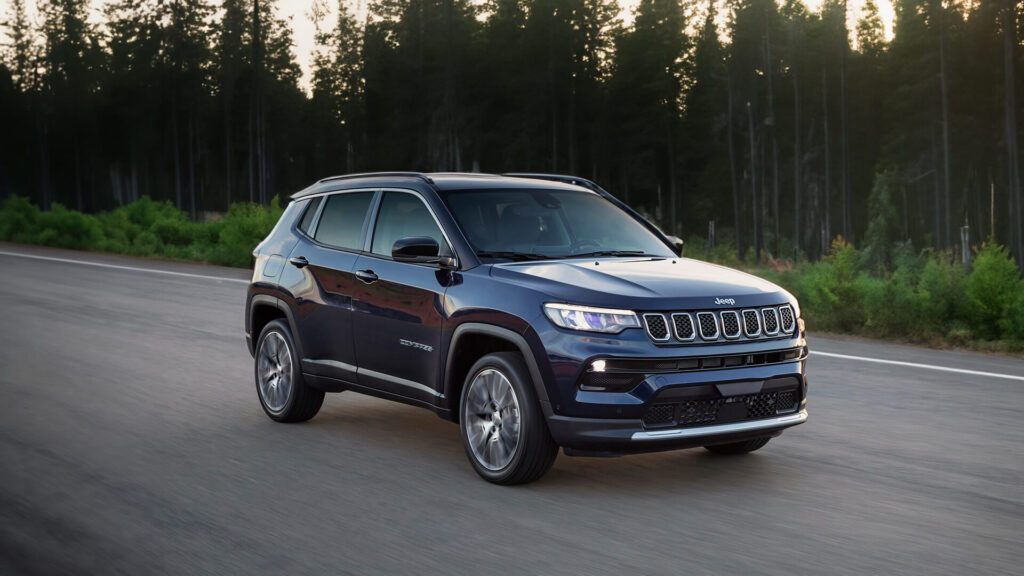 A imagem mostra o SUV Jeep Compass em movimento em uma estrada cercada por árvores altas e densas. O veículo está pintado em uma cor escura, possivelmente preto ou azul marinho, e apresenta a grade de sete fendas característica dos veículos Jeep. Os faróis estão acesos, sugerindo que a foto foi tirada no início da manhã ou no final da tarde. As rodas estão ligeiramente desfocadas, indicando movimento.