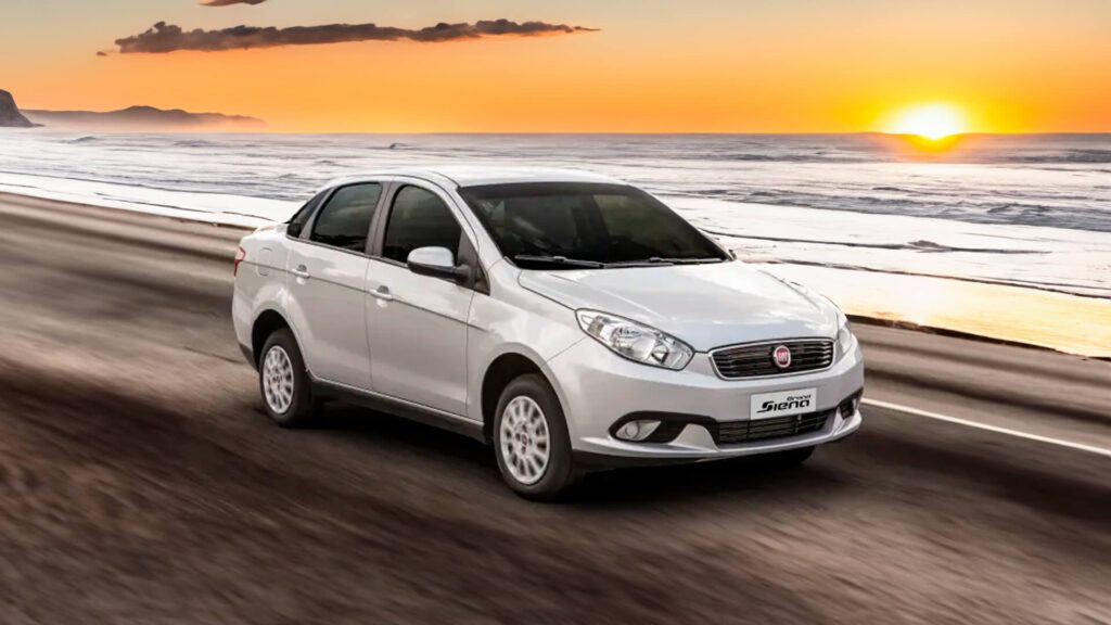 A imagem mostra um carro branco de quatro portas dirigindo ao longo de uma estrada costeira ao pôr do sol. O carro está em movimento, como indicado pelo borrão de movimento na estrada e nas rodas. O fundo apresenta um oceano à esquerda, com ondas suavemente batendo na praia, e um céu claro com tons de laranja e amarelo do sol se pondo.