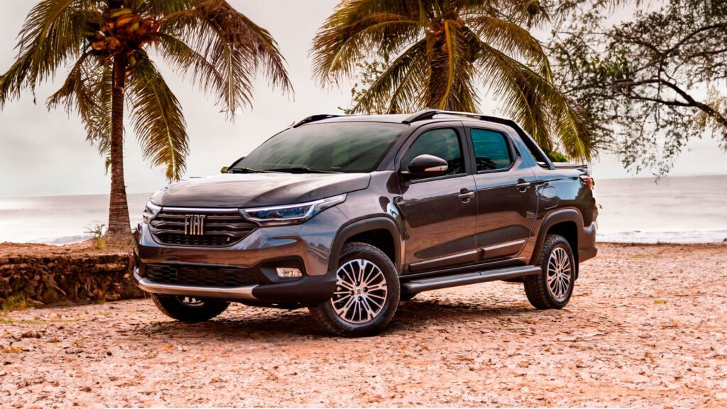 A imagem mostra uma caminhonete Fiat Strada estacionada em uma área de areia com palmeiras ao fundo e um corpo d’água, possivelmente o oceano, visível atrás das árvores. O veículo está posicionado em um ângulo para a câmera, exibindo seu perfil frontal e lateral. Ele tem um design elegante, com uma grade frontal proeminente, linhas contornadas ao longo da carroceria e rodas de liga leve de vários raios. O cenário sugere uma localização tropical ou de praia