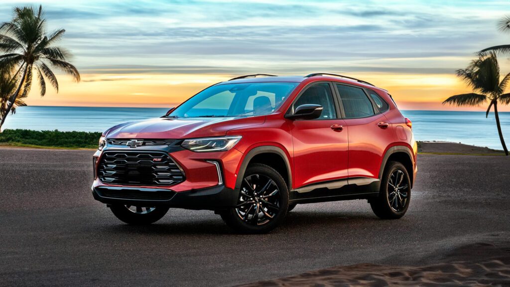 A imagem mostra um SUV Chervrolet Tracker cor vermelho estacionado em uma estrada próxima a uma praia, com o oceano e um pôr do sol ao fundo. O veículo possui detalhes pretos ao redor dos arcos das rodas e rodas pretas, indicando que pode ser um modelo mais esportivo ou off-road. O cenário sugere associação com lazer, viagem ou aventura.