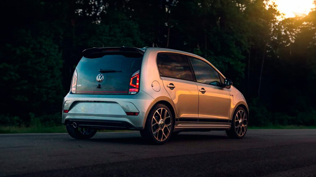 A imagem mostra um carro hatchback prata da Volkswagen estacionado ao lado de uma estrada, com árvores ao fundo. O veículo é capturado em uma vista traseira três quartos durante o que parece ser o pôr do sol, que lança um brilho quente na cena.