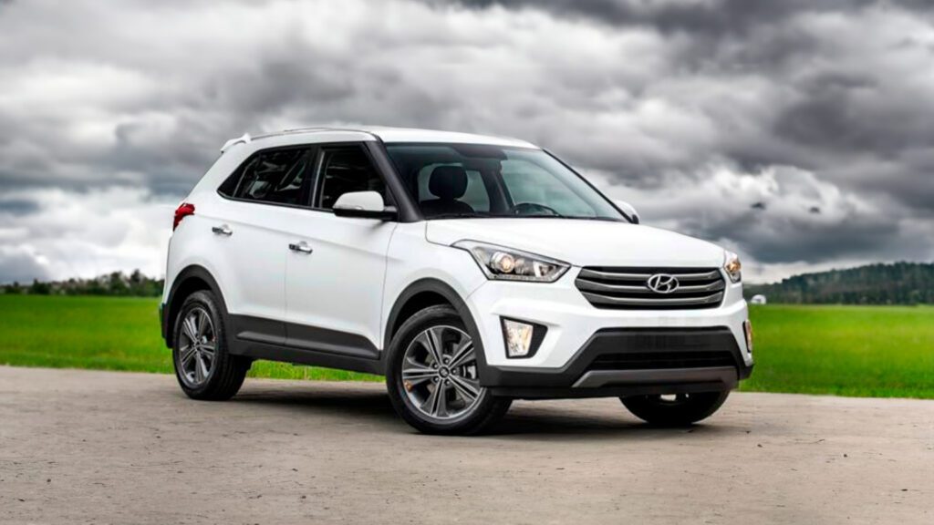 Imagem publicitária para o blog sobre o seguro creta. A imagem mostra um Hyundai Creta branco estacionado em uma superfície de asfalto, com um campo gramado e um céu nublado ao fundo. O veículo está posicionado em um ângulo que destaca seu perfil frontal e lateral, evidenciando características como a grade frontal proeminente, faróis elegantes, faróis de neblina, rodas de liga leve com múltiplos raios e o design compacto de SUV.