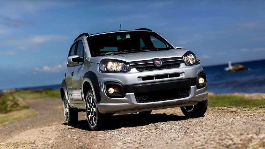 A imagem mostra um Fiat Uno prateado estacionado em uma estrada costeira, com o oceano e um céu azul claro ao fundo. O carro está posicionado em um ângulo que destaca seu perfil frontal e lateral. O logotipo da Fiat é visível na grade frontal, e há um veleiro desfocado ao longe na água.
