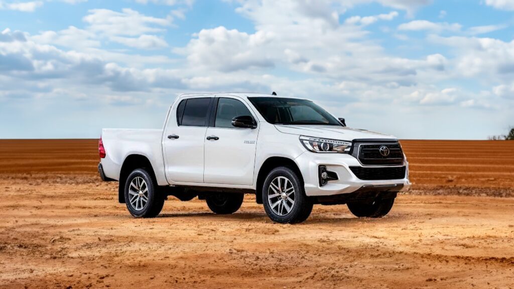 Imagem publicitaria para o blog sobre seguro hilux. A imagem mostra uma Toyota Hilux branca estacionada em uma paisagem plana e árida, com um céu claro ao fundo. O veículo está posicionado em um ângulo que destaca seu perfil frontal e lateral. A Hilux parece ser um modelo mais novo, indicado por suas características de design moderno, como faróis elegantes e grade cromada. O cenário sugere uma capacidade off-road ou uso rural do veículo, o que pode ser relevante para aqueles interessados no desempenho e na estética de picapes em ambientes ao ar livre.