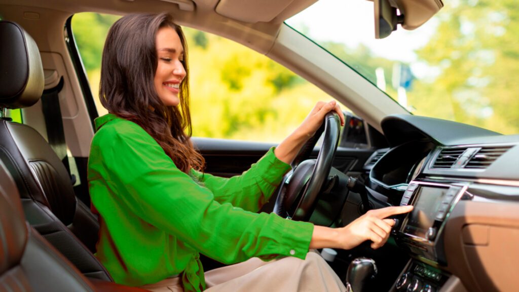 Faça uma cotação de Seguro Auto para mulheres na Natal Seguros. A imagem mostra uma mulher de cabelo castanho dirigindo um carro, com uma das mãos no volante e a outra mão na tela multimídia do carro. Isso sugere que a motorista está interagindo com o sistema de entretenimento ou navegação do veículo. O interior do carro é visível, incluindo o painel, o console central e parte do assento do passageiro. Fora do veículo, há folhagem verde visível através das janelas, indicando que o carro está em uma estrada cercada por vegetação.