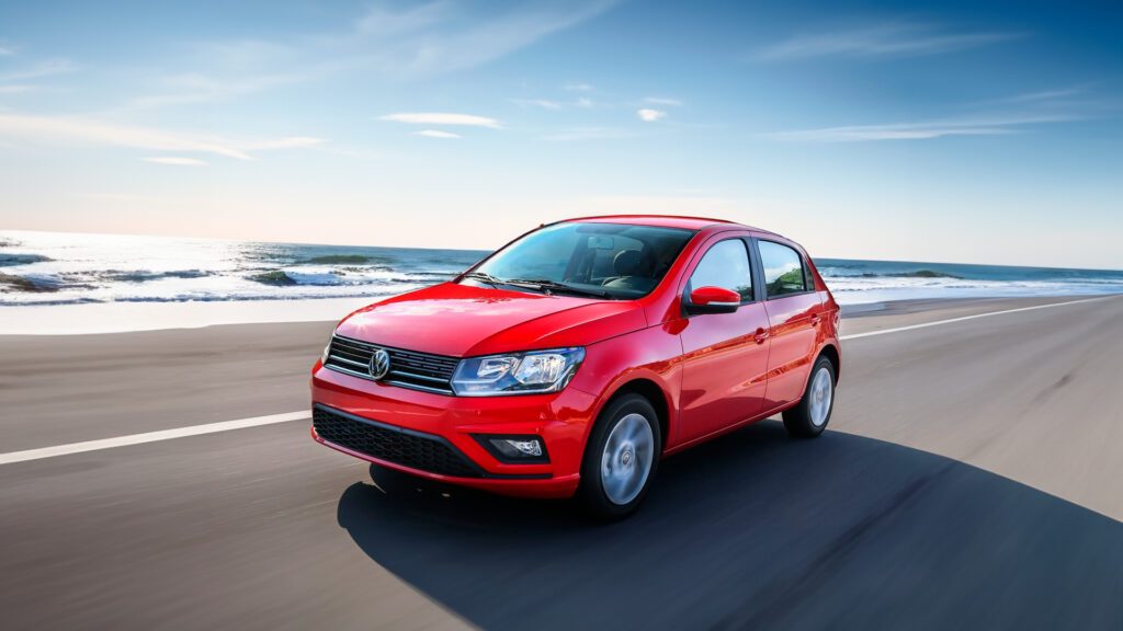 Imagem para o blog post "Seguro do Volkswagen Gol". A imagem mostra um Volkswagen Gol vermelho em movimento, dirigindo em uma estrada com o oceano e ondas visíveis ao fundo. O carro está em movimento, indicado pelo leve desfoque da paisagem e das rodas. O céu está claro com poucas nuvens, sugerindo um clima agradável para dirigir. Esta imagem pode ser interessante para entusiastas da marca Volkswagen, especialmente do modelo Gol, ou para aqueles interessados em fotografia automotiva que captura veículos em movimento dentro de paisagens naturais.