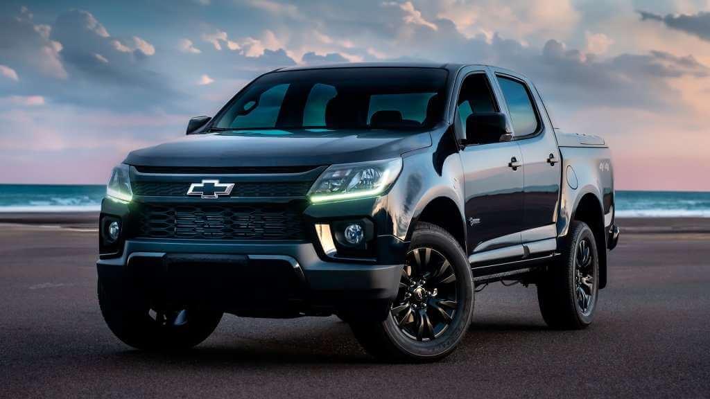 A imagem mostra uma caminhonete Chevrolet S10 preta estacionada em uma praia, com o oceano e um pôr do sol ao fundo. O veículo apresenta um design moderno com linhas elegantes e possui o logotipo da Chevrolet na grade frontal. As rodas têm aros pretos, e os faróis estão apagados. A cena é bastante pitoresca, destacando a caminhonete em um ambiente natural e relaxante.