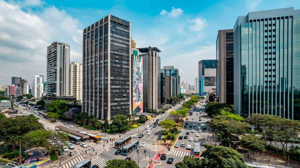 A imagem mostra uma avenida movimentada na cidade de São Paulo, cercada por edifícios altos de diferentes estilos arquitetônicos. No centro, há um grande mural pintado na lateral de um prédio, adicionando um toque vibrante de cor à paisagem urbana. O tráfego flui em várias faixas, com carros e ônibus visíveis. O céu está parcialmente nublado, mas permite que a luz do sol ilumine a cena. Esta imagem captura a natureza dinâmica da vida na cidade de São Paulo e destaca sua mistura de modernidade com expressão artística.