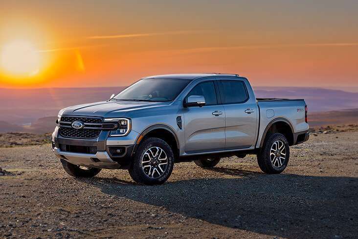 A imagem mostra uma Ford Ranger estacionada em um terreno rochoso com o sol se pondo ao fundo. O veículo está posicionado à esquerda da imagem, capturado em um ângulo lateral-frontal que destaca características como a grade, os faróis e as rodas. O logotipo da Ford é visível na grade frontal e “Ranger” está escrito na parte inferior das portas dianteiras. O pôr do sol proporciona uma luz de fundo quente, criando um contraste interessante com os tons frios da cor do caminhão. Esta imagem parece ser ideal para mostrar o design e o apelo deste modelo específico da Ford Ranger em um cenário ao ar livre, enfatizando suas capacidades como veículo off-road.