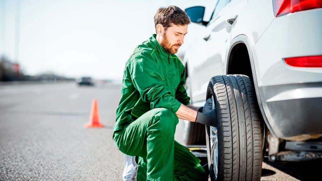 Imagem publicitária para o blog post : Alinhamento e Balanceamento : Qual é a Diferença?. A imagem mostra um homem de jaqueta e calça verde agachada ao lado da traseira de um carro branco, aparentemente trocando um pneu. No fundo, há cones de trânsito laranja, sugerindo que essa atividade está ocorrendo perto de uma estrada ativa ou em um ambiente controlado para segurança. Esta imagem pode ser relevante para discussões sobre manutenção de veículos, procedimentos de segurança na estrada ou como uma ilustração de uma situação cotidiana de resolução de problemas.