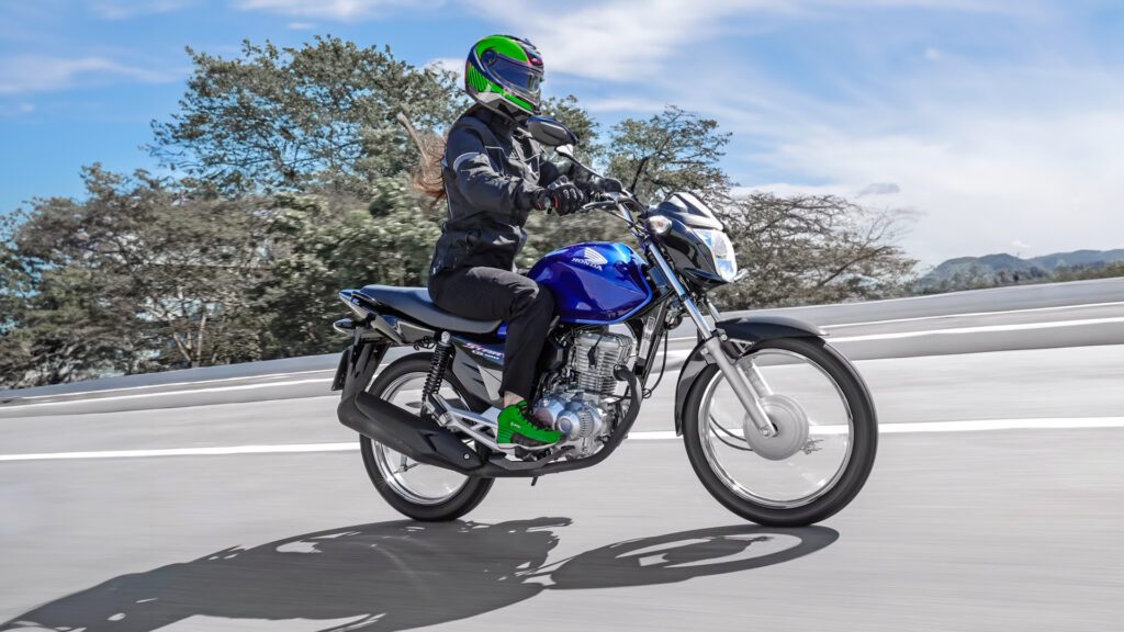 A imagem mostra uma motocicleta Honda CG 160 em movimento, em uma estrada aberta com o céu claro ao fundo. A moto é predominantemente preta com detalhes azuis no tanque de combustível. O piloto está usando um capacete integral com detalhes verdes e roxos, jaqueta preta, luvas e calças escuras. O fundo está desfocado devido ao movimento da motocicleta, destacando a sensação de velocidade.