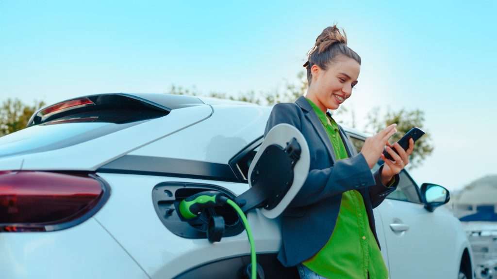 Imagem publicitária para o blog post sobre seguro para carros elétricos. A imagem mostra uma mulher ao lado de um carro elétrico branco que está sendo carregado. O cabo de carregamento está conectado à porta de carregamento do carro. A pessoa está de costas para a câmera, olhando para um smartphone em suas mãos. Esta imagem representa a tecnologia de transporte moderno e a transição para fontes de energia sustentáveis no dia a dia.