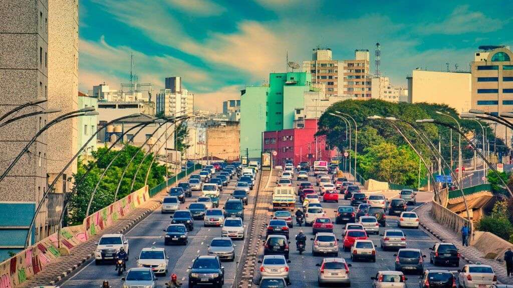 A imagem mostra uma rua urbana movimentada em São Paulo com várias faixas de tráfego. Há muitos carros, sugerindo um congestionamento ou tráfego intenso. A arquitetura dos edifícios ao longo da rua é moderna, com designs e cores variados. Acima, há estruturas que parecem ser parte de uma ponte ou linha de trem elevada. O céu está parcialmente nublado, e a iluminação geral sugere que pode ser fim de tarde ou início da noite. Esta imagem captura a agitação da vida na cidade e pode ser relevante para discussões sobre planejamento urbano, transporte ou gestão de tráfego.