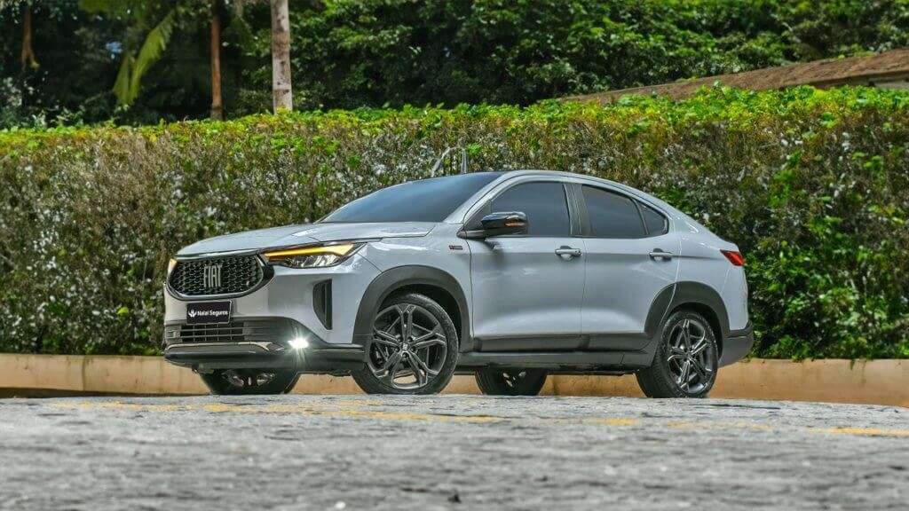 A imagem mostra um carro Fiat Fastback em um ambiente externo. O veículo é de cor prata e está estacionado em uma superfície pavimentada. O design do carro é moderno e esportivo, com linhas aerodinâmicas e rodas de liga leve pretas. A frente do carro apresenta uma grade com o logotipo da Fiat e faróis de LED. O fundo da imagem é composto por uma cerca viva e árvores, sugerindo que o local é uma área verde ou um jardim. A imagem destaca o design elegante e contemporâneo do Fiat Fastback, um modelo que combina características de SUV e coupé, atraindo atenção tanto pela estética quanto pela funcionalidade.