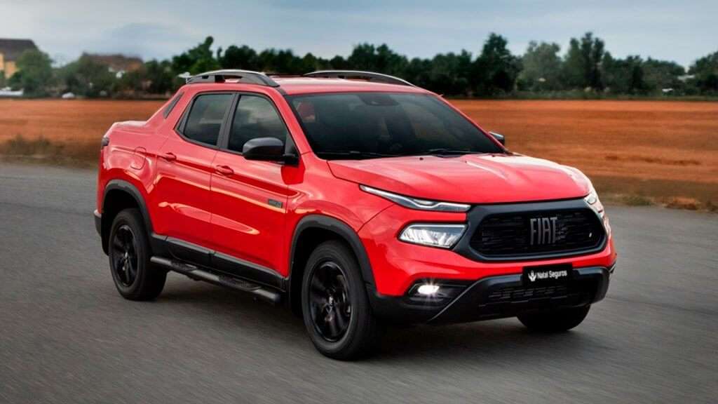 A imagem mostra uma Fiat Toro, um veículo utilitário esportivo (SUV) de porte médio, em um tom vermelho vibrante. A Toro está em movimento em uma estrada asfaltada, com um campo aberto ao fundo.