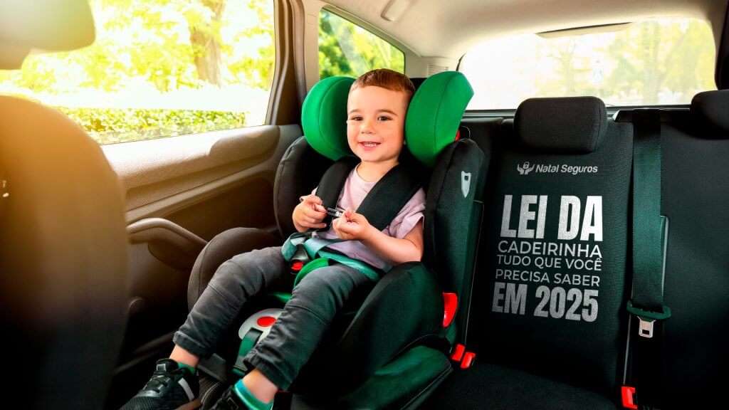 A imagem mostra o interior de um veículo com foco em uma cadeirinha de segurança infantil. Há uma criança sentada na cadeirinha. A cadeirinha é preta com detalhes verdes e parece estar instalada de forma segura no carro, usando um cinto de segurança para manter a criança no lugar. No lado direito da imagem, há um texto que diz “LEI DA CADEIRINHA TUDO QUE VOCÊ PRECISA SABER EM 2025” sugerindo que a imagem pode estar relacionada a novas regulamentações ou leis sobre cadeirinhas de segurança infantil que entraram em vigor em 2024.