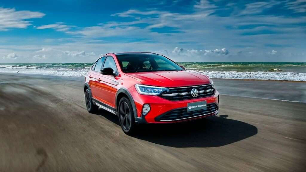 A imagem mostra um Volkswagen Nivus vermelho, um SUV compacto, dirigindo em uma praia com areia clara e mar ao fundo. O carro está em movimento, com as rodas dianteiras levemente viradas para a direita, sugerindo uma curva suave. O céu está azul e claro, com algumas nuvens brancas. A iluminação é natural, com o sol brilhando intensamente, criando um contraste agradável entre o carro vermelho e a paisagem.