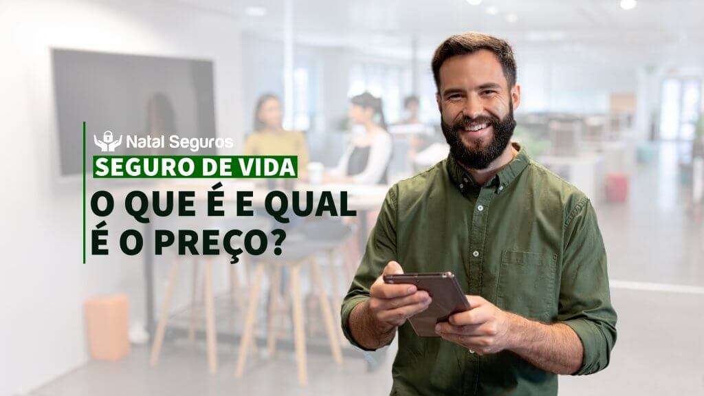A imagem mostra um homem de camisa verde segurando um celular em um ambiente de escritório.Ao fundo, há outras pessoas desfocadas, sentadas em uma mesa de reunião. No lado esquerdo da imagem, há um quadro ou tela desfocada. O texto na imagem diz: Natal Seguros SEGURO DE VIDA O QUE É E QUAL É O PREÇO?