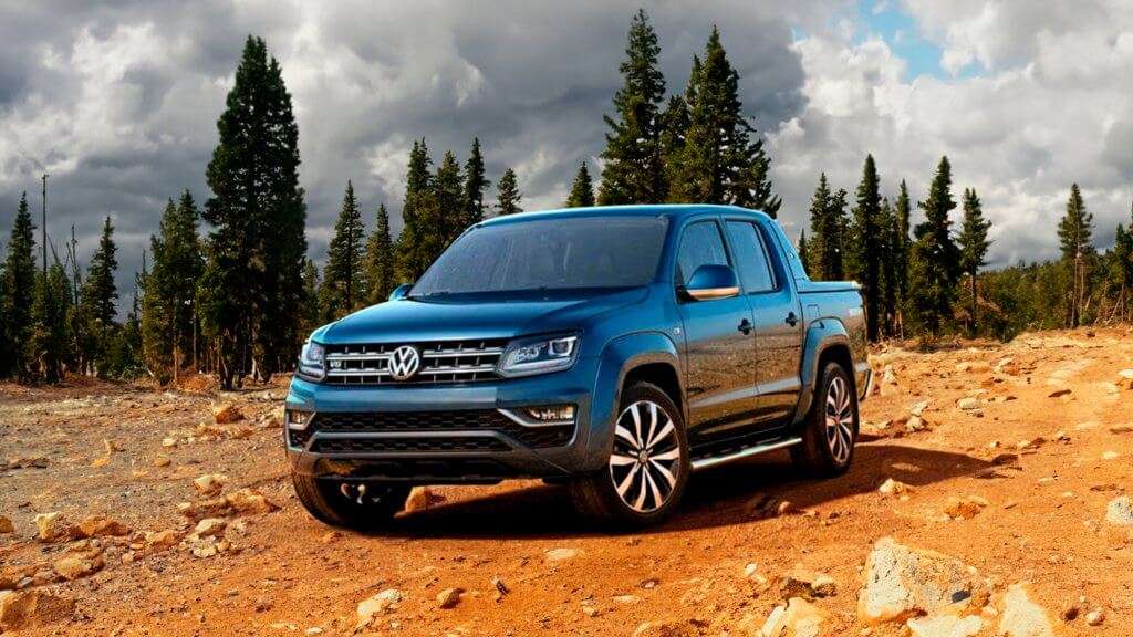 Imagem de capa para o blog sobre seguro amarok. A imagem mostra uma caminhonete Volkswagen Amarok de cor azul, estacionada em um terreno rochoso e arenoso, cercado por uma floresta de pinheiros. O céu está parcialmente nublado, com nuvens escuras e algumas áreas de céu azul visíveis. A caminhonete está posicionada de forma que a frente e o lado direito do veículo são claramente visíveis. A grade frontal exibe o logotipo da Volkswagen, e os faróis estão acesos. As rodas têm um design moderno e esportivo. A imagem destaca a robustez e a capacidade off-road da Amarok, sugerindo que o veículo é adequado para terrenos difíceis e aventuras ao ar livre.