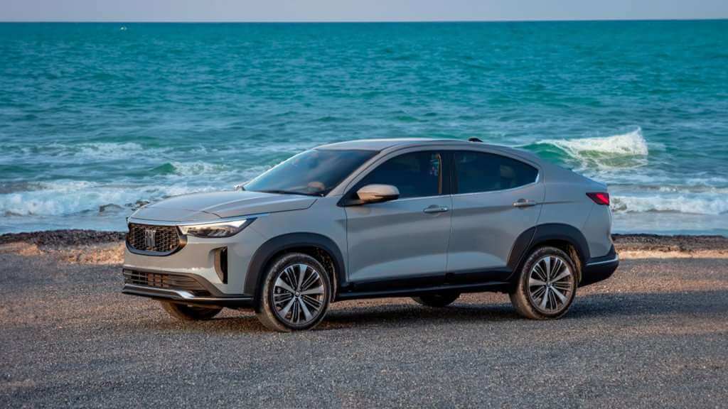 A imagem mostra um carro Fiat Fastback estacionado em uma praia com o mar ao fundo. O veículo é de cor prata e possui um design moderno e aerodinâmico, com linhas suaves e elegantes. As rodas são grandes e possuem um design de raios múltiplos. O cenário ao fundo inclui ondas quebrando na praia e um céu claro, sugerindo um dia ensolarado. A imagem destaca o carro em um ambiente natural, contrastando a engenharia automotiva com a beleza natural do mar.