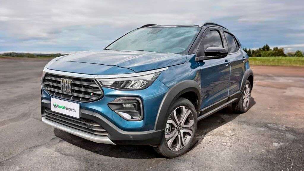 A imagem mostra um Fiat Pulse de cor azul estacionado em um local aberto, possivelmente um estacionamento ou uma estrada. O carro é da marca Fiat, como indicado pelo logotipo na grade frontal. A placa do carro exibe o nome "Natal Seguros". O veículo possui um design moderno com faróis de LED, rodas de liga leve e detalhes cromados.