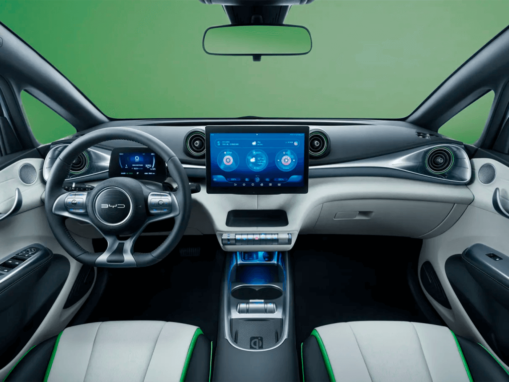 A imagem mostra o interior de um carro BYD Dolphin. O design é moderno e futurista, com um painel de instrumentos digital atrás do volante e uma grande tela sensível ao toque no centro do painel. O volante tem o logotipo da BYD e botões integrados. O painel central exibe informações sobre o veículo e controles de mídia. As saídas de ar têm um design circular e estilizado. O interior é predominantemente em tons de cinza e preto, com detalhes em verde nas bordas dos assentos. O console central possui porta-copos e um compartimento de armazenamento.