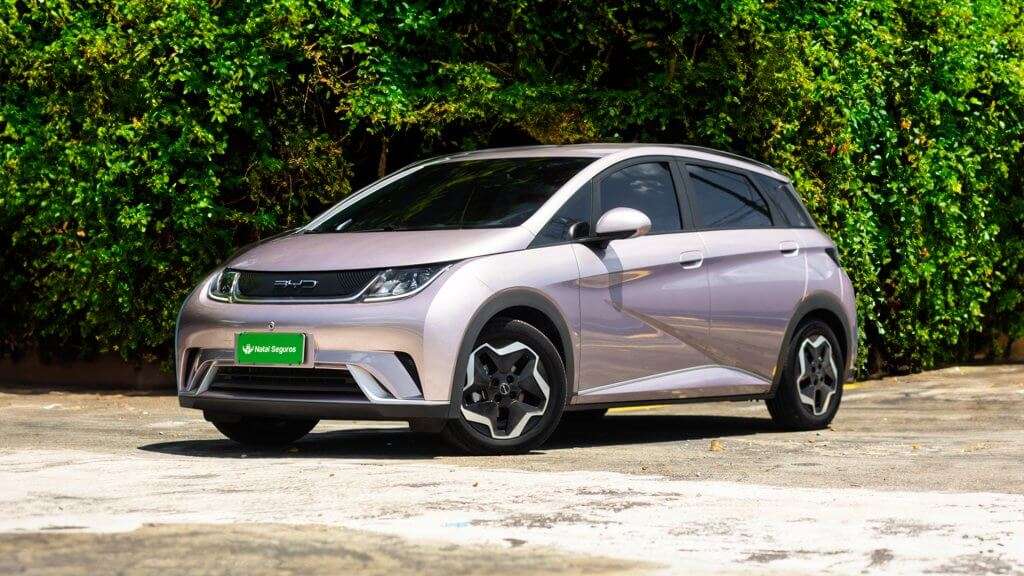 A imagem mostra um carro elétrico BYD Dolphin EV estacionado em uma área ao ar livre, com uma vegetação verde ao fundo. O carro tem uma cor metálica clara e um design moderno e aerodinâmico. As rodas possuem um design distinto com detalhes em preto e prata. A placa do carro é verde e exibe o texto "Natal Seguros". O BYD Dolphin EV é interessante por ser um veículo elétrico, representando uma opção sustentável e inovadora no mercado automotivo.