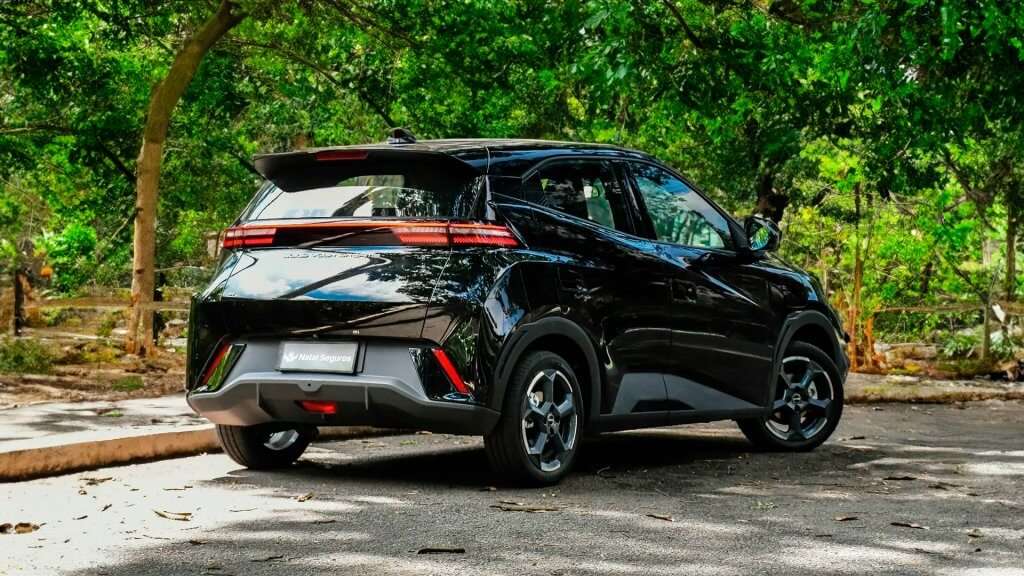 A imagem mostra a traseira de um carro preto estacionado em um ambiente arborizado. O carro é um BYD Dolphin Mini, um veículo elétrico compacto. A traseira do carro apresenta um design moderno com linhas angulares e luzes traseiras horizontais que se estendem por toda a largura do veículo. A placa do carro tem a inscrição "Natal Seguros". O ambiente ao redor é verde e natural, com árvores e vegetação densa. O carro está estacionado em uma área pavimentada, possivelmente em um parque ou área de lazer.