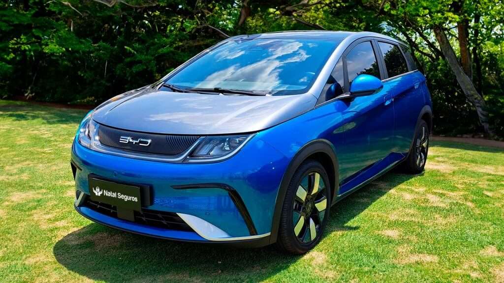 A imagem mostra um carro elétrico azul da marca BYD, modelo Dolphin Plus, estacionado em um gramado verde. O carro possui um design moderno e aerodinâmico, com faróis de LED e rodas de liga leve. Na frente do veículo, há uma placa preta com o texto "Natal Seguros". O logotipo da BYD está visível na grade frontal do carro. Ao fundo, há uma vegetação densa com árvores e arbustos, sugerindo que o carro está em um ambiente natural.