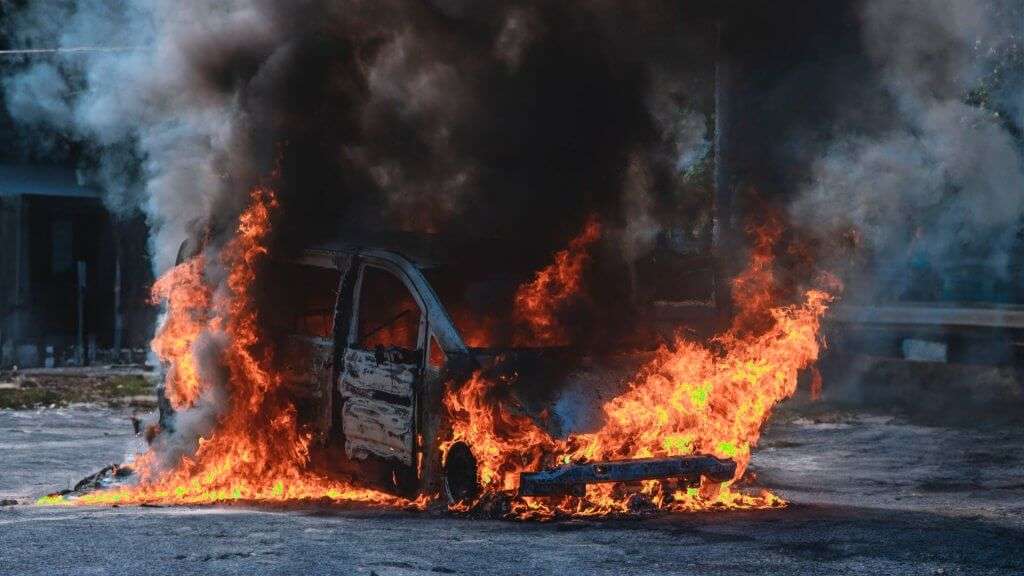 A imagem mostra um carro em chamas, com grandes labaredas de fogo e uma densa fumaça preta subindo ao céu. O veículo parece estar completamente envolvido pelo fogo, com chamas intensas saindo de várias partes do carro. A cena é dramática e chama a atenção devido à intensidade do incêndio e à quantidade de fumaça gerada. Não há pessoas visíveis na imagem, e o cenário ao redor parece ser uma área urbana ou suburbana. A imagem é relevante por destacar um incidente de incêndio veicular, que pode ser de interesse para questões de segurança, investigação de causas de incêndio, ou simplesmente como um registro visual impactante de um evento perigoso.