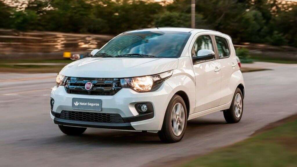 A imagem mostra um carro Fiat Mobi branco em movimento em uma estrada. O carro está em destaque, com os faróis acesos e a placa frontal exibindo o nome "Natal Seguros". O fundo está desfocado, sugerindo que o carro está em movimento. O Fiat Mobi é um carro compacto, popular em áreas urbanas devido ao seu tamanho reduzido e eficiência de combustível.