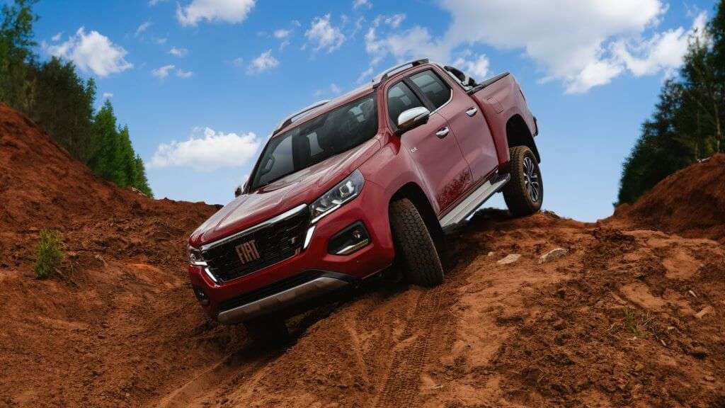 A imagem mostra uma caminhonete Fiat Titano de cor vermelha em um terreno acidentado de terra. O veículo está inclinado, sugerindo que está em uma situação de off-road, demonstrando sua capacidade de enfrentar terrenos difíceis. O céu está azul com algumas nuvens brancas, e há árvores verdes ao fundo, indicando que a cena ocorre em uma área natural. A caminhonete parece robusta e adequada para aventuras fora de estrada, o que pode ser interessante para entusiastas de veículos 4x4 e off-road.