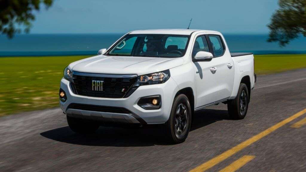 A imagem mostra uma caminhonete Fiat Titano Volcano branca em movimento em uma estrada. O veículo está em destaque, com o logotipo da Fiat visível na grade frontal. Ao fundo, há uma paisagem com gramado verde e o mar ao longe, sob um céu azul claro. A imagem é interessante e relevante por destacar o design moderno e robusto da caminhonete, além de sugerir um ambiente de viagem ou aventura.