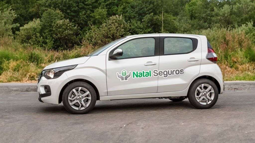 A imagem mostra um carro Fiat Mobi branco estacionado em uma estrada pavimentada com vegetação ao fundo. Na lateral do carro, há um adesivo com o logotipo e o nome "Natal Seguros". O logotipo consiste em um ícone de cadeado verde dentro de um círculo, com duas mãos estilizadas em cinza segurando o círculo. O texto "Natal Seguros" está escrito ao lado do logotipo, com "Natal" em verde e "Seguros" em cinza.