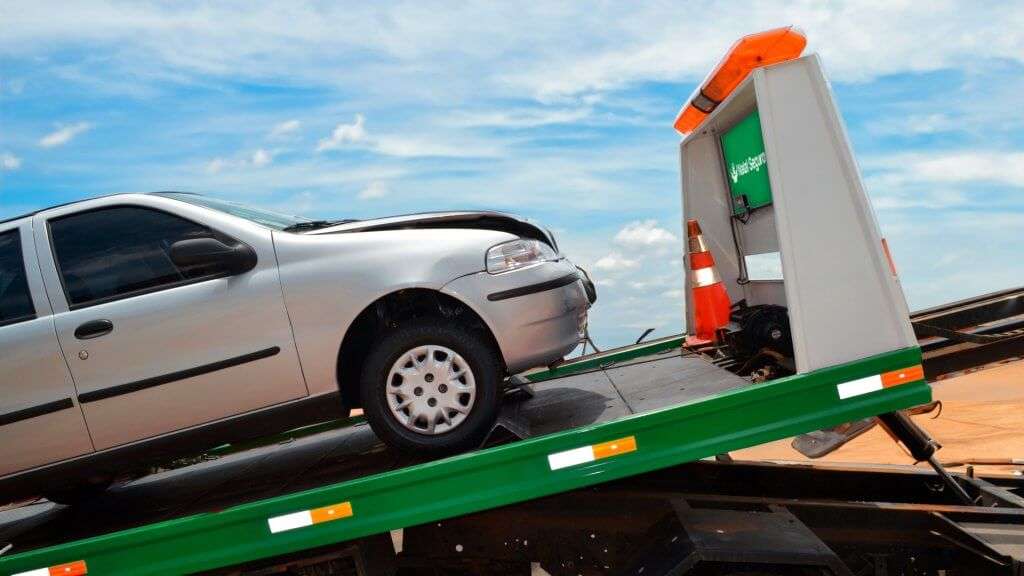 A imagem mostra um carro Fiat Palio de cor prata sendo rebocado por um guincho. O carro está inclinado sobre a plataforma do guincho, indicando que foi carregado recentemente. A frente do carro está danificada, sugerindo que esteve envolvido em um acidente. No guincho, há um cone de sinalização laranja e uma luz de emergência laranja no topo. O céu está claro com algumas nuvens, indicando que é um dia ensolarado.