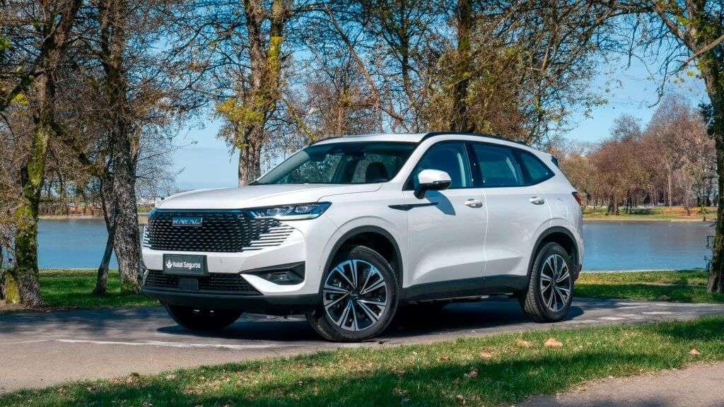 A imagem mostra um carro SUV branco, especificamente um Haval H6 PHEV (Plug-in Hybrid Electric Vehicle), estacionado em uma área arborizada próxima a um lago. O carro está posicionado de forma que a frente e o lado direito estejam visíveis.  O veículo possui um design moderno e elegante, com uma grade frontal grande e faróis de LED. As rodas têm um design esportivo, e os retrovisores são brancos, combinando com a cor do carro. No para-choque dianteiro, há uma placa com o nome "Natal Seguros".  Ao fundo, é possível ver árvores com poucas folhas e um corpo d'água, sugerindo que a foto foi tirada em um parque ou área natural. A imagem destaca o design e a presença de um veículo híbrido plug-in, que é uma opção mais sustentável e eficiente em termos de consumo de combustível.