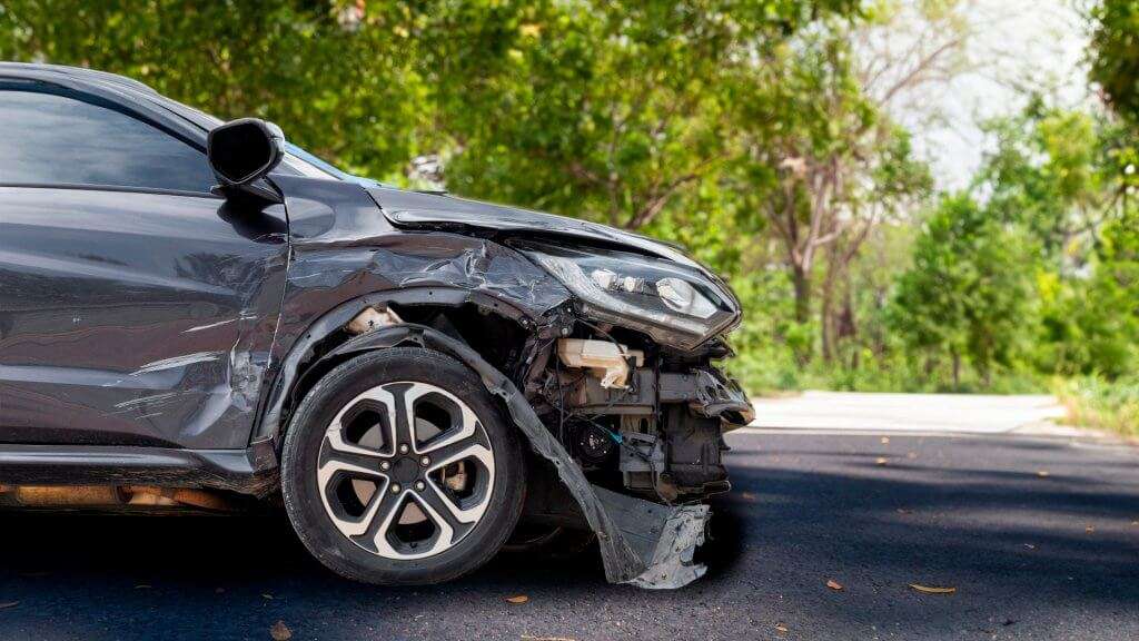 A imagem mostra um Honda HRV após uma colisão. O veículo está visivelmente danificado na parte frontal direita, com o para-choque, o farol e a área ao redor da roda dianteira direita severamente amassados e quebrados. Partes internas do carro, como componentes mecânicos e elétricos, estão expostas devido ao impacto. A imagem foi tirada em uma estrada asfaltada com árvores e vegetação ao fundo, sugerindo que o acidente pode ter ocorrido em uma área arborizada ou em uma estrada rural. A relevância da imagem está em ilustrar os danos significativos que um veículo pode sofrer em uma colisão, destacando a importância da segurança no trânsito e da manutenção adequada dos veículos.