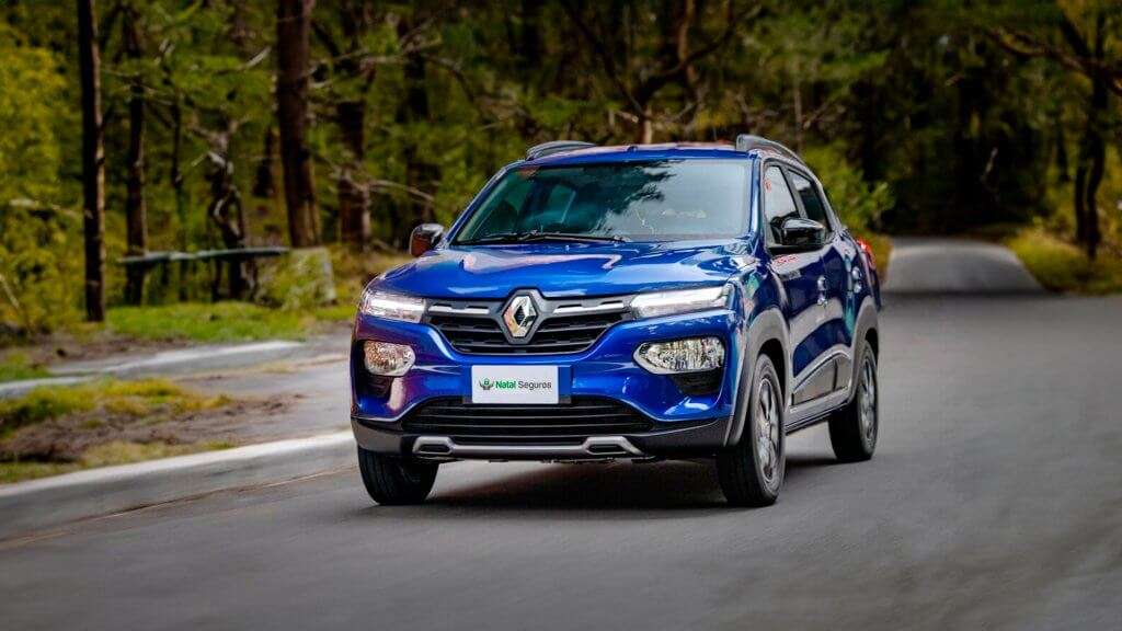 A imagem mostra um carro Renault Kwid 2024 de cor azul, em movimento em uma estrada cercada por árvores. O carro está em destaque no centro da imagem, com a frente voltada para a câmera. A estrada parece ser uma via rural ou uma estrada em meio a uma floresta, com vegetação verde ao redor. O design moderno e robusto do Renault Kwid 2024 é bem evidente, destacando-se no ambiente natural e tranquilo.