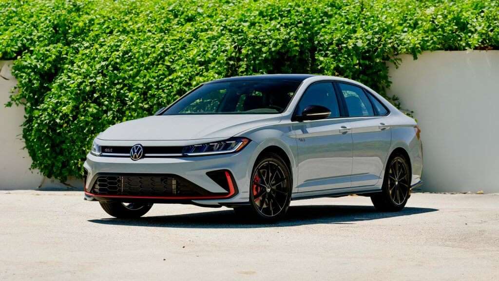 A imagem mostra um Volkswagen Jetta GLI estacionado em uma área externa, com um fundo de vegetação verde e um muro branco. O carro é de cor cinza claro, com detalhes em preto e vermelho, incluindo rodas pretas com pinças de freio vermelhas. O emblema "GLI" está visível na grade frontal e na lateral do veículo. O design do carro é esportivo e moderno, com linhas elegantes e faróis de LED.