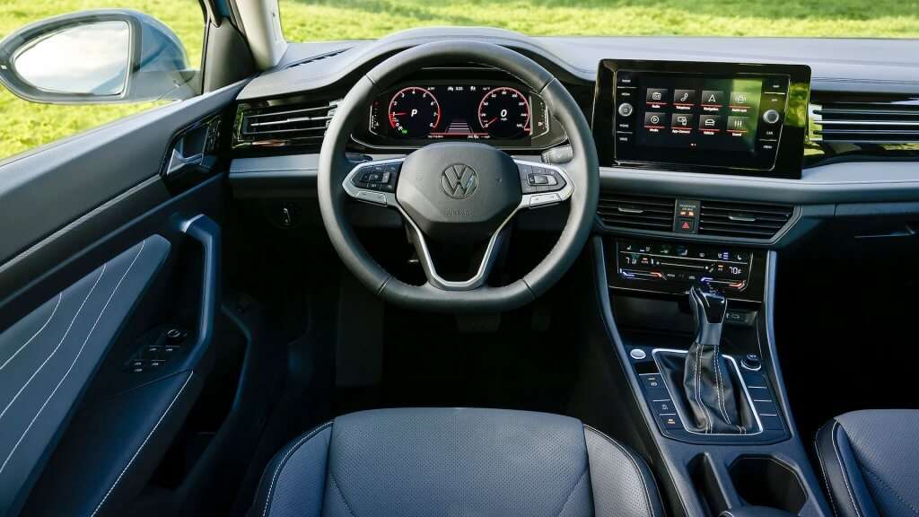  Descreva essa imagem (Interior Jetta GLI) A imagem mostra o interior de um Volkswagen Jetta GLI. O volante está centralizado com o logotipo da Volkswagen no meio e botões multifuncionais nas laterais. Atrás do volante, há um painel de instrumentos digital com dois mostradores circulares e várias informações exibidas